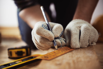 Close-up of craftsman hands in protective gloves measuring wooden plank with ruler and pencil. Woodwork and renovation concept.  - obrazy, fototapety, plakaty