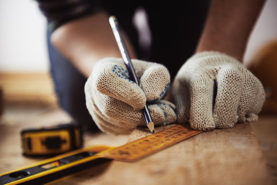 Close-up of craftsman hands in protective gloves measuring wooden plank with ruler and pencil. Woodwork and renovation concept. 