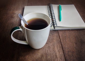Cup of hot black coffee with background of blank note and pen on wood table in vintage instagram colour look for concept of creation accompany with coffee