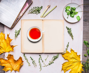 cup of tea standing on an open book with autumn leaves, books, mint and thyme on wooden rustic background top view
