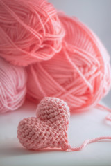 Adorable little heart crocheted by hand. Made with a thread of a thick bulky wool thread. Three skeins of baby pink color yarns are visible on the background..