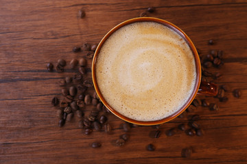 Brown ceramic cup of coffee decorated with roasted beans around on wooden background, close up