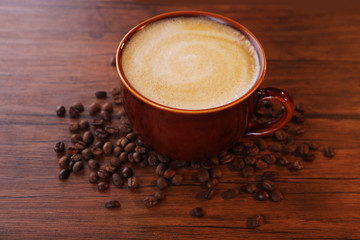 Brown ceramic cup of coffee decorated with roasted beans around on wooden background, close up