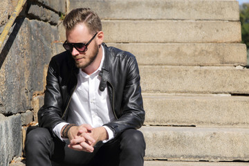 A handsome man in his 20s sitting down outside on a sunny summer day wearing a white shirt and black jeans with a leather jacket. The river behind him.