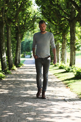 A lonely man wearing a sweater and jeans, walking along a footpath/alley on a sunny summer day, with a focused look on his face.