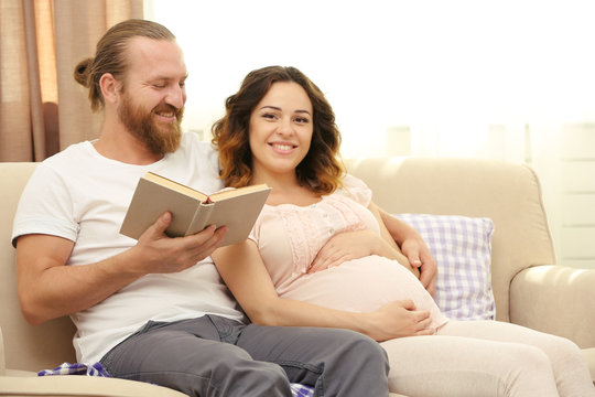 Handsome man reads book to his lovely pregnant woman on sofa in the room
