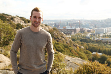 Trendy good looking man standing high up on a mountain with a great view of the city behind him on a sunny day.