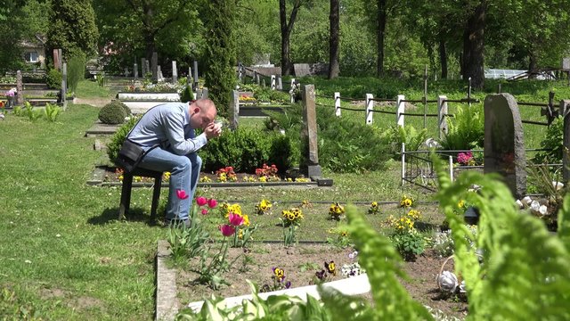 Painful widower man sit shrinked on bench near mother wife tomb and pray in graveyard. Static shot. 4K
