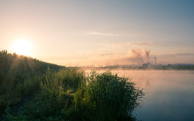 Fototapeta na wymiar Beautiful early morning mist over a lake and a factory pipe polluting air in the distance. Environmental problems background