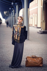 Young lonely sad blonde girl in a coat, gloves, a scarf and a skirt with a short haircut is waiting on a deserted train station with two leather bags, autumn