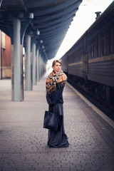 Fototapeta na wymiar Young lonely sad blonde girl in a coat, gloves, a scarf and a skirt with a short haircut is waiting on a deserted train station with two leather bags, autumn