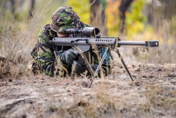 Sniper at work/Man in military uniform lyi on ground in forest and aim through the optical sight