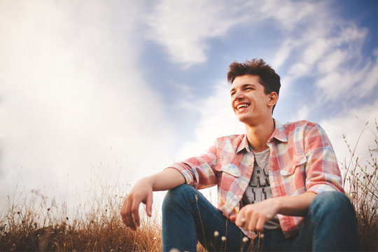 Handsome young man sitting and smiling on sky background. Attractive caucasian teenage boy wearing shirt laughing and looking confident with copy space.