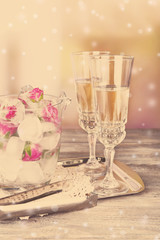 Ice cubes with rose flowers in glass bucket and two glasses with champagne on wooden table, on bright background