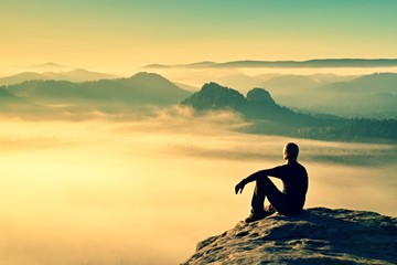 Hiker in black on the rocky peak. Wonderful daybreak in mountains, heavy orange mist in valley. Man sit on the rock.