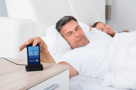 Man On Bed Snoozing Alarm Clock On Cell Phone