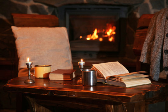 Hot tea or coffee in mug, book and candles on vintage wood table. Fireplace as background