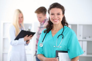 Beautiful young smiling female doctor standing at hospital with doctor and patient on the background. Medical concept.