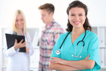 Fototapeta premium Beautiful young smiling female doctor standing at hospital with doctor and patient on the background. Medical concept.