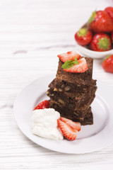 Stack of square pieces of chocolate brownies with hazelnuts, served with whipped cream, strawberry and mint on white wooden table. Delicious breakfast concept. Space for text. Selective focus