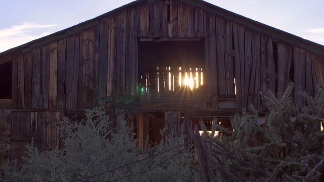 Old wooden abandoned brown barn with sun gleaming and streaming through open slats and beams in the dilapidated wood with sagebrush and plants in foreground with slow dolly move.