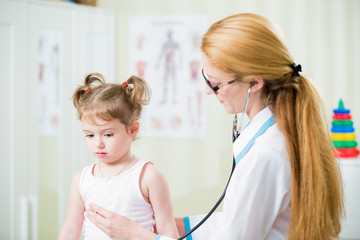 Pediatrician woman examining cute little girl with stethoscope. Kid looks sad and sick
