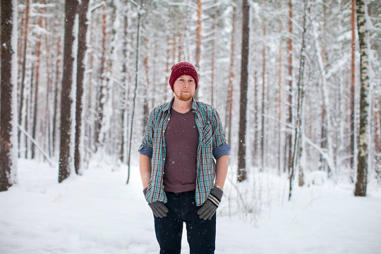 a man with a beard wearing a shirt and hat standing in a winter forest