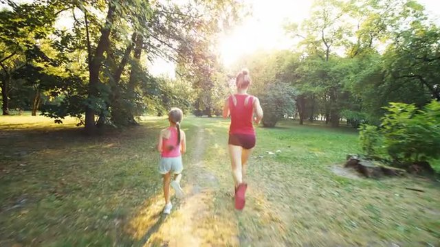 Athletes family mother and daughter running away from camera in the park in sunlight. Lens flare