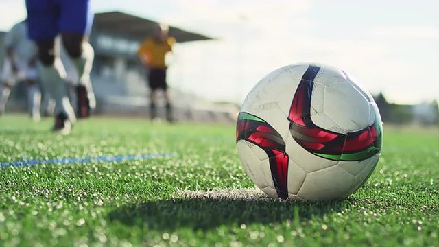 Close up of a soccer ball being kicked by a player in slow motion, with players in the background

