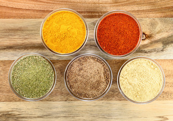 The powders of: bay leaf, turmeric, mustard, red paprika and nutmeg are poured in the glass bowls on the wooden background.
