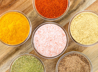 The powders of: bay leaf, turmeric, mustard, red paprika and nutmeg are poured in the glass bowls on the wooden background.