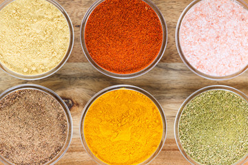 The powders of: bay leaf, turmeric, mustard, red paprika and nutmeg are poured in the glass bowls on the wooden background.