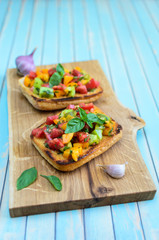 Fresh homemade rustic bruschetta with chopped red, yellow and green tomatoes, basil, olive oil and toasted garlic italian bread ciabatta on wooden cutting board over turquoise background close-up