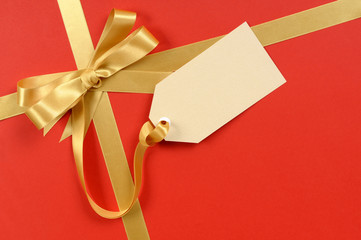 Gold bow ribbon decoration with plain gift tag on red wrapping paper background for christmas or...