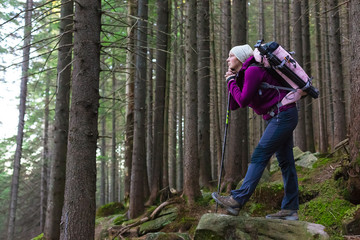 Female Hiker Staying inside Deep Old Forest on Moss Stone with Backpack and Trekking Pole Looking into Distance with Pensive Face