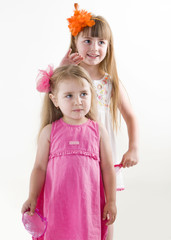  The older sister on the white dress takes care of the younger on the pink dress combing her hair and gets lipstick on her lips isolated on the white background