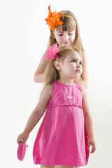  The older sister on the white dress takes care of the younger on the pink dress combing her hair and gets lipstick on her lips isolated on the white background
