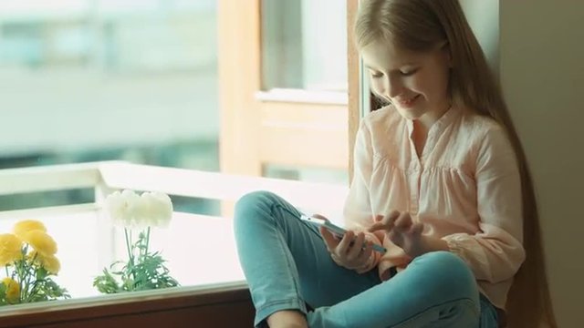 Child using mobile phone and smiling. Looking at camera. Shows ok. Thumbs up. Girl sitting on the window sill