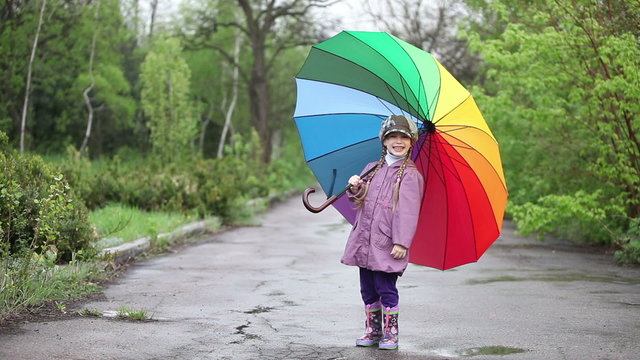 Cheerful little girl hiding behind umbrella in the park. Thumbs up. Waving hand at the camera