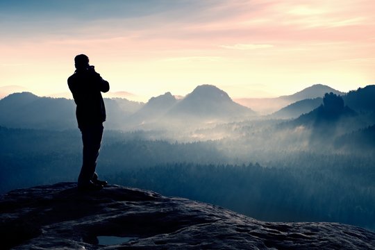 Professional photographer takes photos with big camera on rock. Dangerous possition at end of cliff.  Dreamy  foggy landscape, hot Sun above