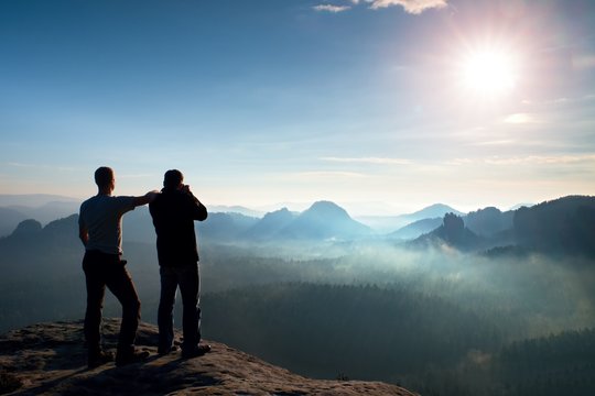 Two friends. Hiker thinking and photo enthusiast takes photos  stay on cliff. Dreamy fogy landscape, blue misty sunrise in a beautiful valley below