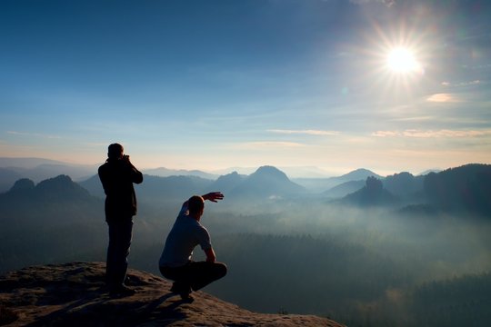 Two friends. Hiker thinking and photo enthusiast takes photos  stay on cliff. Dreamy fogy landscape, blue misty sunrise in a beautiful valley below