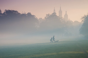 Blonia meadow in Krakow, Poland, with St Mary's church and Town Hall towers in the background, foggy morning with people's silhouettes