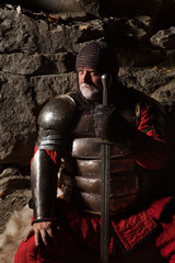Fototapeta na wymiar Old medieval King in armor with sword is sitting on furs
