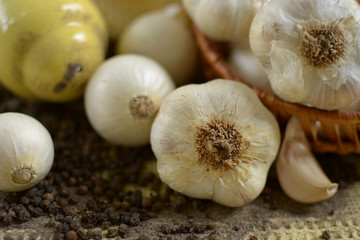 garlic in front of basket with garlics on white ground pepper and black pepper corns, dipped yellow pepper pot on yellow tablecloth