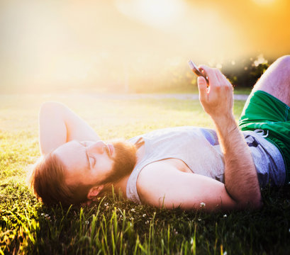 young man with a beard and brown hair, a T-shirt and shorts, lying on the grass in the sunlight with a smartphone in hand