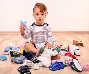 Toddler playing with a lot of baby shoes holding one shoe up. Untidy stack of child shoes thrown on the ground.