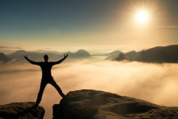 Fototapeta premium Funny jumping hiker in black celebrate triumph. Stay between two rocky peaks. Wonderful misty daybreak.