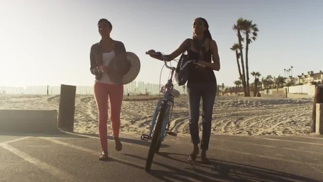 Two African American women friends walking bike through parking lot at the beach at sunset