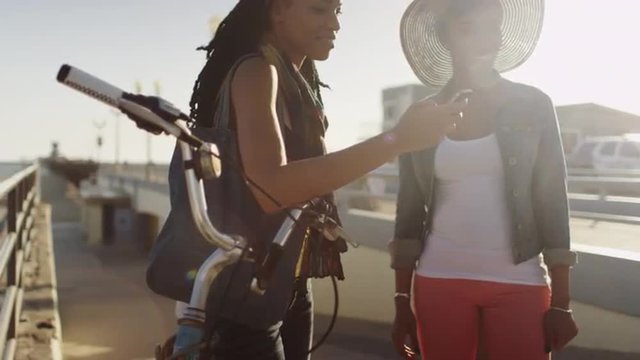 Backlit handheld shot of two black women friends texting and taking pictures on the pier